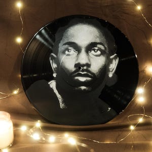 May include: Black and white vinyl record featuring a portrait of a man. The record is illuminated by string lights and a lit candle in a glass jar. The background is a dark brown color.