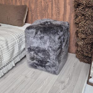 May include: A square, gray faux fur ottoman. The ottoman is placed on a light gray wood floor next to a bed with a white blanket and a brown pillow. The ottoman is a cube shape and has a soft, fluffy texture.
