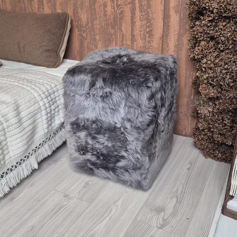 May include: A square, gray faux fur ottoman. The ottoman is placed on a light gray wood floor next to a bed with a white blanket and a brown pillow. The ottoman is a cube shape and has a soft, fluffy texture.