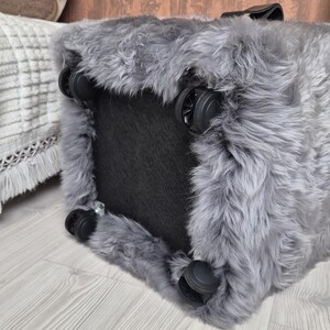 May include: A gray, fluffy storage ottoman with four black wheels for easy movement. The ottoman has a black base and a handle on top. The faux fur material gives it a soft, inviting appearance. The wheels are made of black plastic.