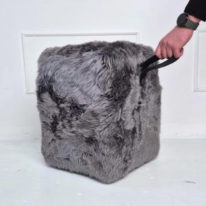 May include: A gray, cube-shaped ottoman with a shaggy, faux fur texture. The ottoman has a black handle on top. The background is a white wall and floor.