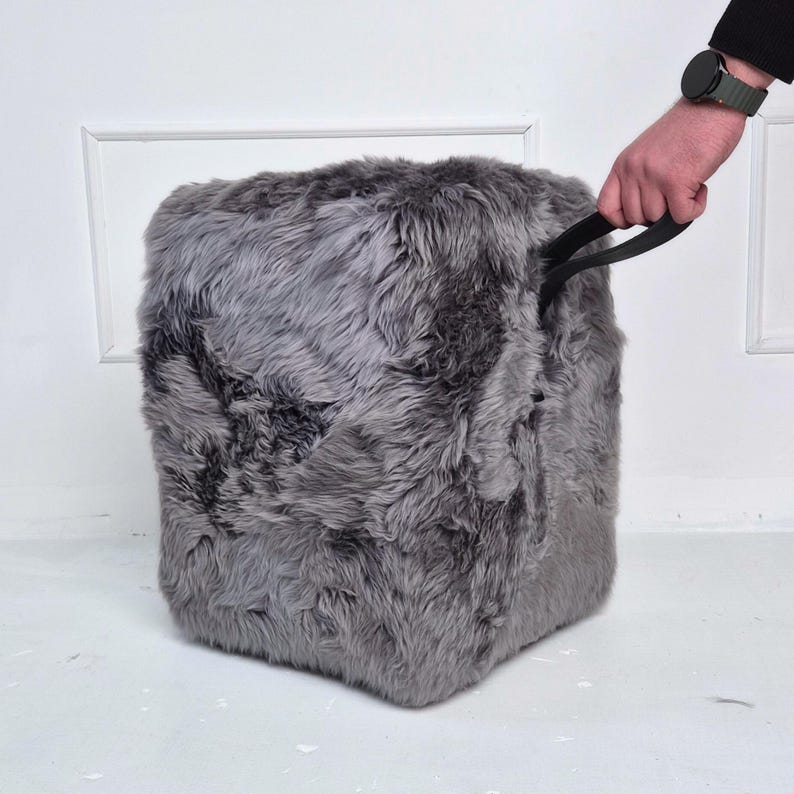 May include: A gray, cube-shaped ottoman with a shaggy, faux fur texture. The ottoman has a black handle on top. The background is a white wall and floor.