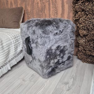 May include: A square, gray faux fur ottoman with a black leather handle. The ottoman is placed on a light wood floor. A bed with a white blanket and brown pillow is in the background.