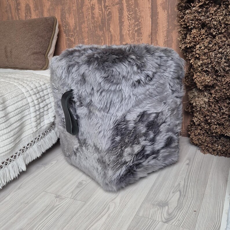 May include: A square, gray faux fur ottoman with a black leather handle. The ottoman is placed on a light wood floor. A bed with a white blanket and brown pillow is in the background.