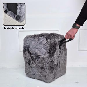 May include: A gray, cube-shaped ottoman with a shaggy, faux fur texture. The ottoman has a black handle and sits on invisible wheels. The image includes the text "Invisible wheels".