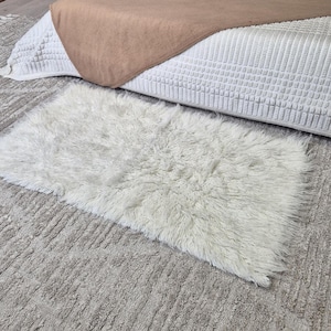 Puede incluir: Una alfombra rectangular, esponjosa y blanquecina con fibras largas se encuentra sobre una alfombra gris texturizada. Una colcha blanca y una manta beige están en el fondo. La alfombra añade una textura suave y acogedora a la habitación.