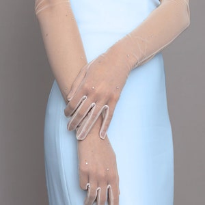 May include: Sheer, beige-toned gloves with sparkling rhinestone accents. The gloves are long, extending up the arms, and are paired with a light blue dress. The gloves have a delicate, elegant appearance, suitable for formal occasions.