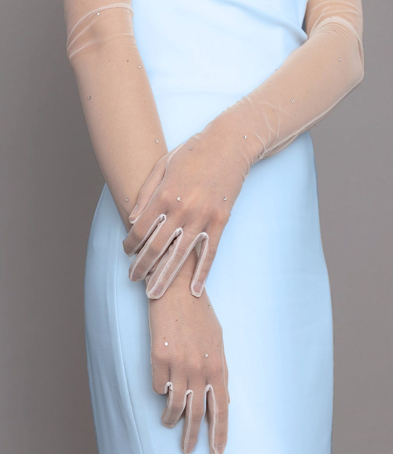 May include: Sheer, beige-toned gloves with sparkling rhinestone accents. The gloves are long, extending up the arms, and are paired with a light blue dress. The gloves have a delicate, elegant appearance, suitable for formal occasions.