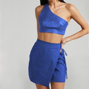 Puede incluir: Conjunto de dos piezas azul cobalto. La parte superior es un top corto de un solo hombro. La parte inferior es una falda envolvente. El conjunto est&aacute; hecho de una tela texturizada. La modelo lleva pendientes dorados.