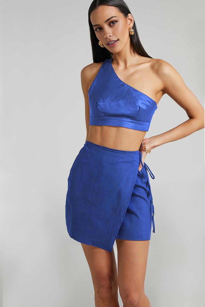 Puede incluir: Conjunto de dos piezas azul cobalto. La parte superior es un top corto de un solo hombro. La parte inferior es una falda envolvente. El conjunto est&aacute; hecho de una tela texturizada. La modelo lleva pendientes dorados.
