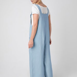 May include: Pale blue linen jumpsuit with a wide leg and square neckline. The jumpsuit has adjustable straps and pockets. A white t-shirt is worn underneath. The outfit is paired with light brown sandals.