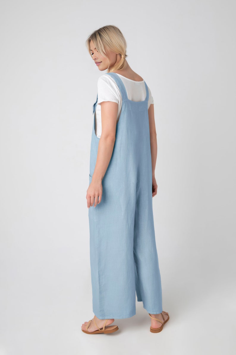May include: Pale blue linen jumpsuit with a wide leg and square neckline. The jumpsuit has adjustable straps and pockets. A white t-shirt is worn underneath. The outfit is paired with light brown sandals.