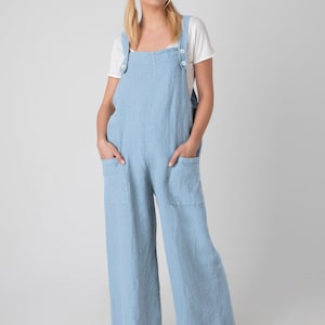 May include: Pale blue linen overalls with large front pockets and button fastenings on the straps. The model wears a white t-shirt. The overalls have wide legs and are worn with sandals.