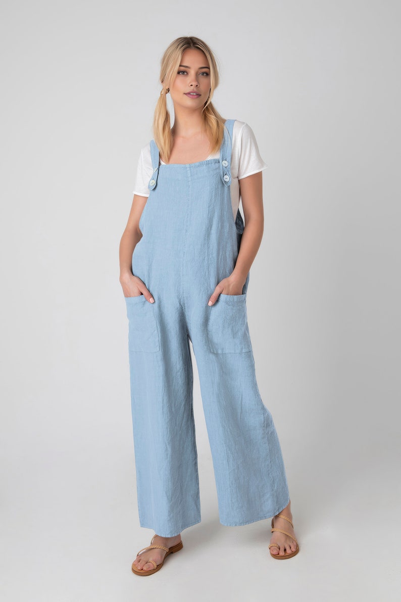 May include: Pale blue linen overalls with large front pockets and button fastenings on the straps. The model wears a white t-shirt. The overalls have wide legs and are worn with sandals.