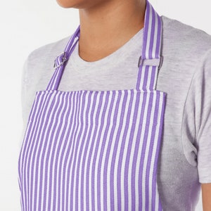 May include: Close-up of a purple and white striped apron. The apron has a bib and adjustable straps with metal hardware. The apron is worn over a gray t-shirt. The stripes are vertical and evenly spaced.