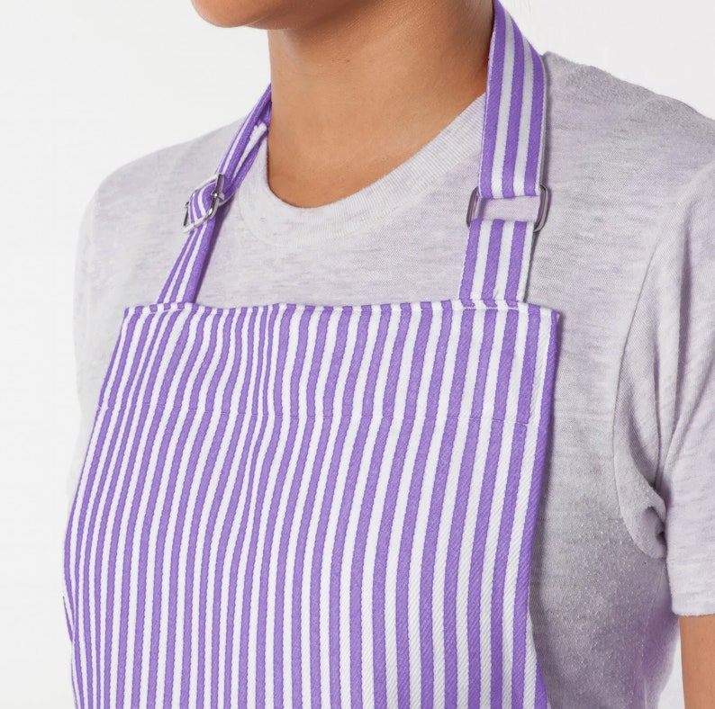 May include: Close-up of a purple and white striped apron. The apron has a bib and adjustable straps with metal hardware. The apron is worn over a gray t-shirt. The stripes are vertical and evenly spaced.