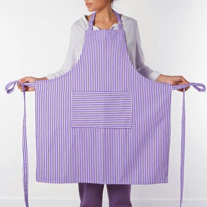 May include: A purple and white striped apron with a large front pocket. The apron has a neck strap and long waist ties. The apron is made of a lightweight fabric.