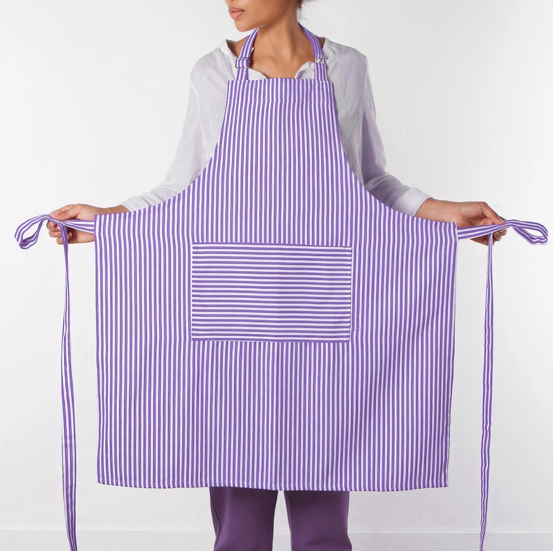 May include: A purple and white striped apron with a large front pocket. The apron has a neck strap and long waist ties. The apron is made of a lightweight fabric.