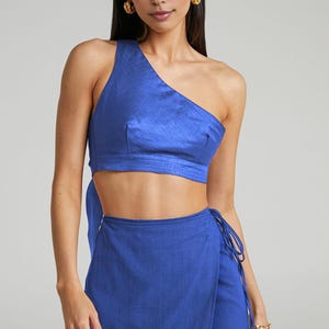 Puede incluir: Conjunto de dos piezas azul cobalto. La parte superior es un top corto de un solo hombro. La parte inferior es una falda envolvente con cierre de lazo. La modelo lleva pendientes dorados y una pulsera dorada.