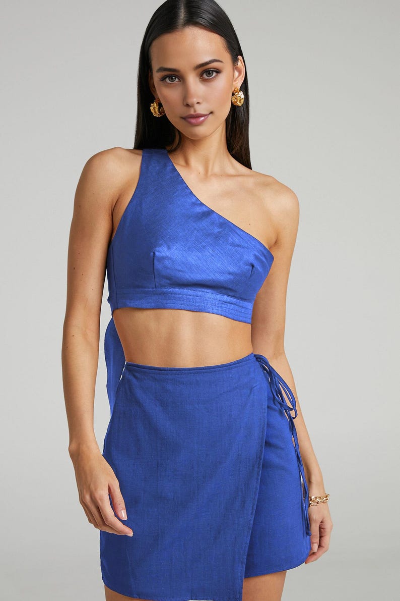 Puede incluir: Conjunto de dos piezas azul cobalto. La parte superior es un top corto de un solo hombro. La parte inferior es una falda envolvente con cierre de lazo. La modelo lleva pendientes dorados y una pulsera dorada.