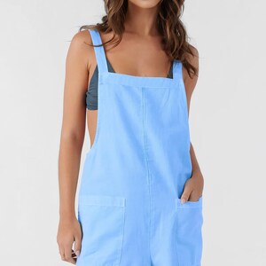 May include: Pale blue romper with a square neckline and broad shoulder straps. It has two front pockets and rolled-up hems. The model is wearing a dark grey top. The style is casual and suitable for summer wear.