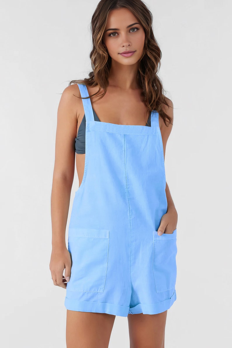 May include: Pale blue romper with a square neckline and broad shoulder straps. It has two front pockets and rolled-up hems. The model is wearing a dark grey top. The style is casual and suitable for summer wear.