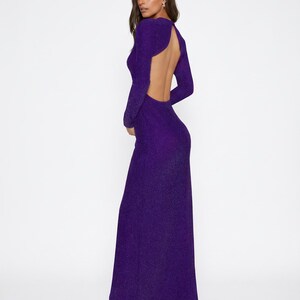 May include: A long, shimmering purple gown with long sleeves and an open back. The dress has a high neckline in the front and a flowing skirt. The fabric appears to have a subtle sparkle, catching the light. The dress is floor-length.