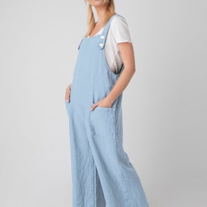 May include: Pale blue linen jumpsuit with wide legs and large front pockets. The jumpsuit has adjustable straps with white buttons and is worn over a white t-shirt. The model is wearing brown sandals.