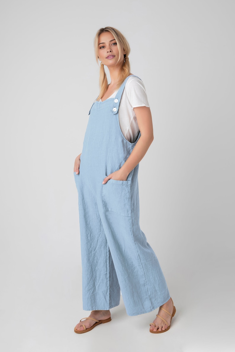 May include: Pale blue linen jumpsuit with wide legs and large front pockets. The jumpsuit has adjustable straps with white buttons and is worn over a white t-shirt. The model is wearing brown sandals.