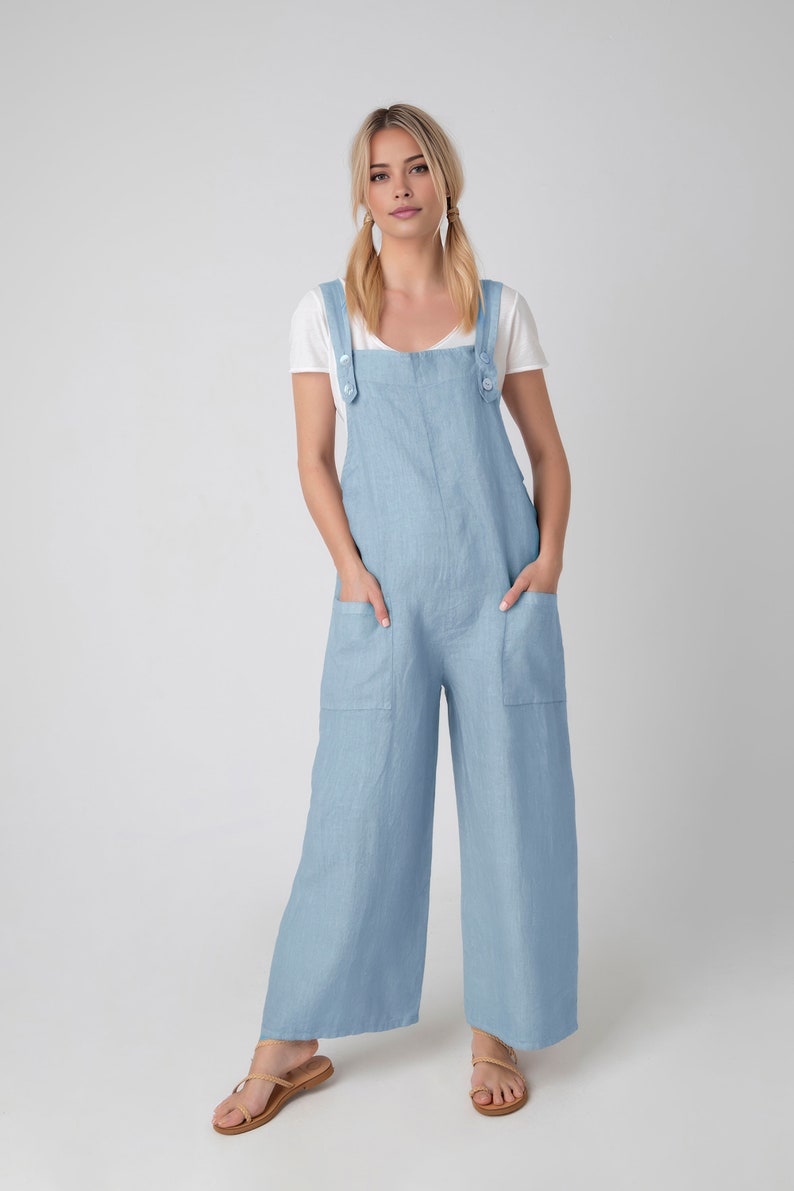May include: Pale blue linen jumpsuit with wide legs and large front pockets. The jumpsuit features adjustable straps with button fastenings and is worn over a white t-shirt. The model is wearing brown sandals.