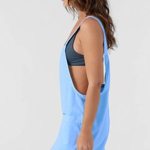 May include: Pale blue romper with a square neckline and rolled cuffs. The romper has a relaxed fit and features two front pockets. The straps are wide and the back is open. The garment is made of a lightweight fabric.