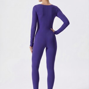 May include: A long-sleeved, form-fitting jumpsuit in a vibrant purple hue. The one-piece garment features a scoop neckline and a streamlined silhouette. The model is wearing white and beige sneakers. The jumpsuit is made of a stretchy material.