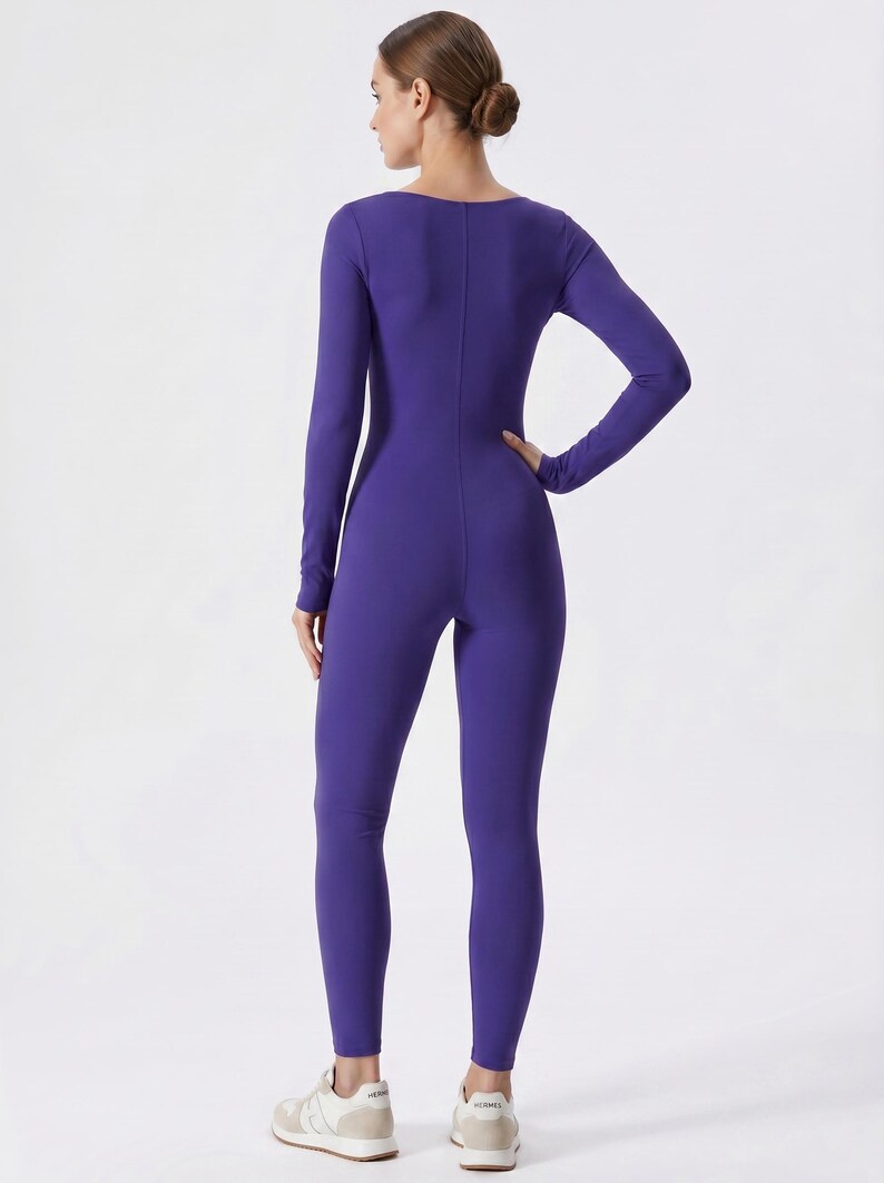 May include: A long-sleeved, form-fitting jumpsuit in a vibrant purple hue. The one-piece garment features a scoop neckline and a streamlined silhouette. The model is wearing white and beige sneakers. The jumpsuit is made of a stretchy material.
