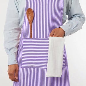 May include: A purple and white striped apron with a large pocket. A wooden spoon is tucked into the apron, and a white kitchen towel is held in the pocket. The apron is worn over a light blue shirt.