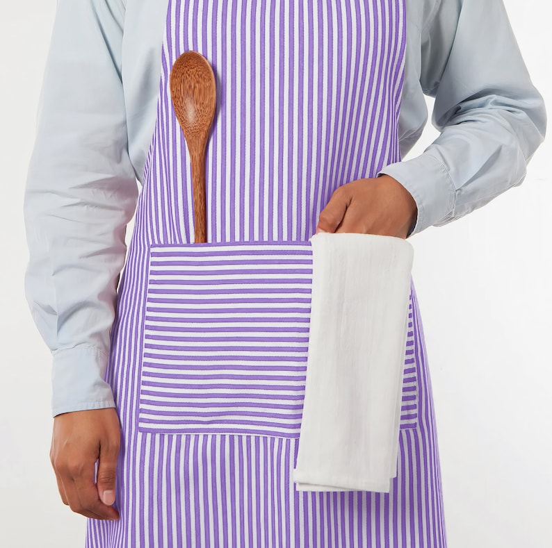 May include: A purple and white striped apron with a large pocket. A wooden spoon is tucked into the apron, and a white kitchen towel is held in the pocket. The apron is worn over a light blue shirt.