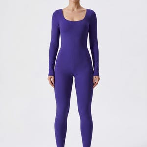 May include: A full-body, form-fitting jumpsuit in a vibrant purple hue. The jumpsuit features a scoop neckline and long sleeves. The model is wearing beige and white sneakers. The overall style is sleek and modern.