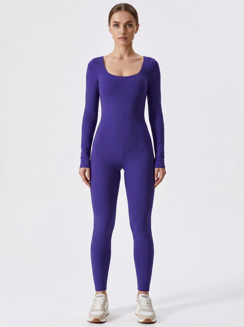 May include: A full-body, form-fitting jumpsuit in a vibrant purple hue. The jumpsuit features a scoop neckline and long sleeves. The model is wearing beige and white sneakers. The overall style is sleek and modern.