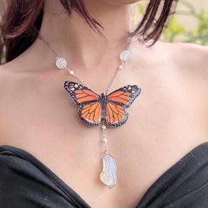 May include: A silver necklace featuring a monarch butterfly pendant with orange and black wings. The necklace includes white beads and a dangling, irregularly shaped, white stone pendant. The butterfly pendant is the focal point of this nature-inspired jewelry.
