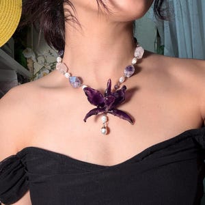 May include: A necklace featuring a large, dark purple flower pendant with a dangling pearl. The necklace also includes a mix of white pearls and irregularly shaped amethyst and rose quartz stones. The necklace is worn on a person.