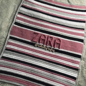 May include: A pink and grey striped crocheted baby blanket with the name "Zara" spelled out in raised pink yarn. The blanket has a black border and the text "@HookedByHayley" is written below the name.