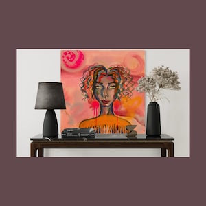 Könnte beinhalten: Ein abstraktes Gemälde einer Frau mit lockigem Haar in Orange-, Rosa- und Blautönen hängt über einem dunklen Holztisch. Eine schwarze Lampe und eine Vase mit getrockneten Blumen stehen auf dem Tisch, zusammen mit Büchern und dekorativen Objekten.