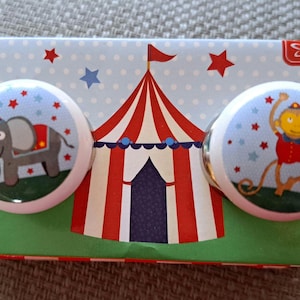 May include: Two ceramic knobs with a circus theme. One knob features a grey elephant wearing a red hat and a red and white striped saddle. The other knob features a yellow monkey wearing a red and white striped shirt and a red hat. Both knobs have a blue background with white stars.