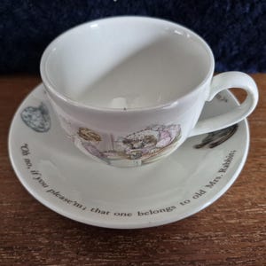 May include: A white teacup and saucer set with a charming illustration and text. The saucer is inscribed with the words "Oh no, if you please'm; that one belongs to old Mrs. Rabbits." The cup has a handle and a matching design.