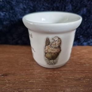 May include: A white ceramic egg cup featuring a detailed illustration of a hen. The egg cup has a rounded base and a wide rim. The hen is depicted in shades of brown, tan, and white. The background is a blurred blue.