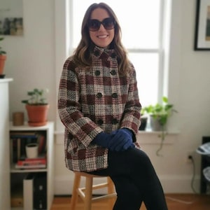 Puede incluir: Una chaqueta de doble botonadura con un patrón de cuadros en marrón, crema y negro, cuello alto y botones oscuros. La chaqueta se combina con guantes azul oscuro lisos y leggings negros, junto con unas gafas de sol grandes de cristales oscuros.