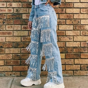 May include: Light blue denim jacket and jeans with silver fringe detailing. The outfit includes a white t-shirt and white sneakers. The background is a brick wall.