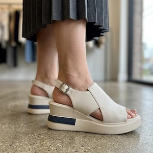 May include: Off-white wedge sandals featuring a wide front strap, an open toe, and an adjustable ankle strap with a silver-toned metal buckle. The platform sole is off-white with a contrasting dark blue rectangular accent on the outer heel.