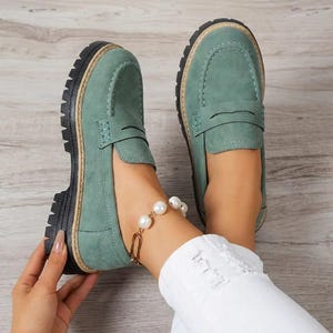 May include: Sage green loafers with a thick black sole and light brown trim. These penny loafers have a classic design with a rounded toe and a strap. The shoes are made of a suede-like material. The shoes are shown with a pearl bracelet.