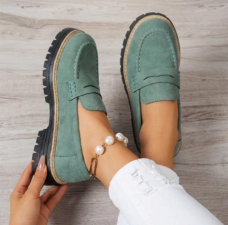 May include: Sage green loafers with a thick black sole and light brown trim. These penny loafers have a classic design with a rounded toe and a strap. The shoes are made of a suede-like material. The shoes are shown with a pearl bracelet.