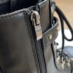 May include: Close-up of a black leather boot with a silver zipper and metal accents. The boot features a lace-up design and a chunky sole, showcasing a modern, edgy style.